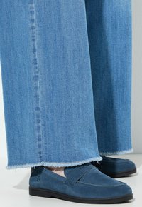 Mocasines de ante azul con un acabado texturizado, combinados con pantalones de mezclilla de pierna ancha, de lavado claro y con dobladillo deshilachado. Suelas negras simples.