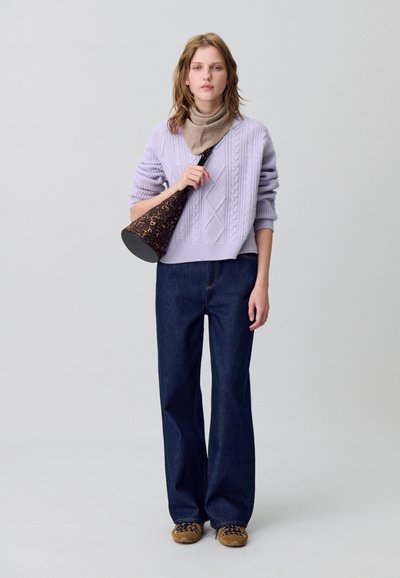 Maglione a maglia cable lilla, sciarpa beige, jeans a gamba larga blu scuro e scarpe stringate marroni. Tiene una borsa a forma di cono con stampa.