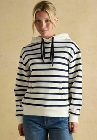 Młoda kobieta o blond włosach, ubrana w białą bluzę z niebieskimi poziomymi paskami i niebieskie dżinsy, stojąca na neutralnym tle.