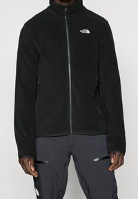 Musta fleece-takki, jossa on korkea kaulus ja täysipitkä vetoketju. Sisältää brodeeratun logon rinnassa ja vetoketjulliset taskut sivuilla.