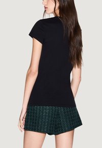 Camiseta negra de manga corta hecha de algodón, combinada con pantalones cortos verdes con estampado. La vista desde atrás muestra el diseño sencillo y el ajuste de la camiseta.