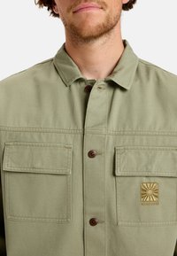 Light green, short-sleeve button-up jacket with two front pockets, brown buttons, and a logo patch on the left pocket. Cotton fabric, plain texture.