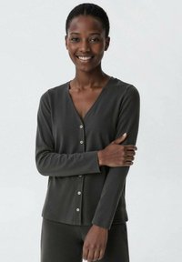 Smiling person wearing a dark buttoned long-sleeve top, standing with one arm crossed and hand resting on the opposite arm.