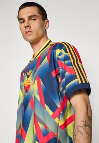 Vícebarevný sportovní dres s geometrickými vzory v modré, žluté, červené a zelené barvě. Obsahuje žlutý límec a tři pruhované rukávy.