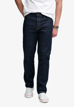 Mann trägt dunkelblaue Jeans, weiße Sneakers, weißes T-Shirt und Armbanduhr, steht mit einer Hand teilweise in der Tasche.