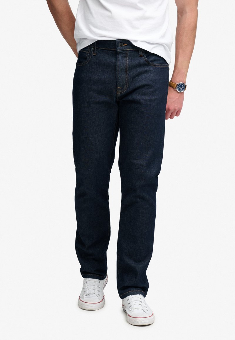 Hombre con vaqueros azul oscuro, zapatillas blancas, camiseta blanca y reloj de pulsera, de pie con una mano parcialmente en el bolsillo.