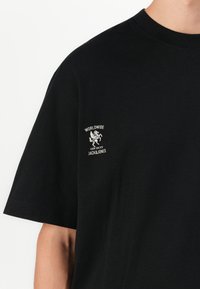 Juoda trumpų rankovių marškinėliai su balta logo ant krūtinės, kuriame pavaizduotas sparnuotas arklys ir tekstas "WORLDWIDE OFF DUTY JACK&JONES."