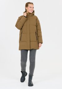 Bruine, gewatteerde parka met een hoge kraag, ritssluitingen en een capuchon. Draagt over grijze jeans en zwarte waterdichte laarzen.