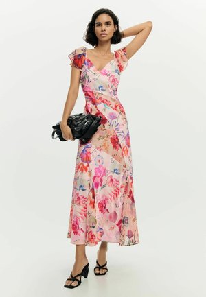 Žena v ružových kvetinových šatách s výstrihom do V, drží čiernu tašku, jednu ruku zdvihnutú, má na sebe čierne sandále na opätku, stojí na bielom pozadí.