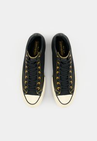 Zapatillas de lona negras con acentos amarillos, que presentan una puntera de goma blanca, cordones negros y detalles de costura en contraste.
