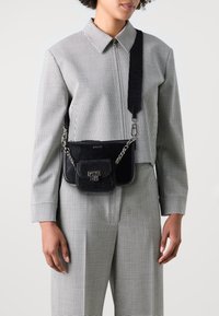 Borsa a tracolla nera in pelle con finitura testurizzata, dettagli in catena e una piccola tasca frontale. Indossata con una camicia a scacchi e pantaloni coordinati.