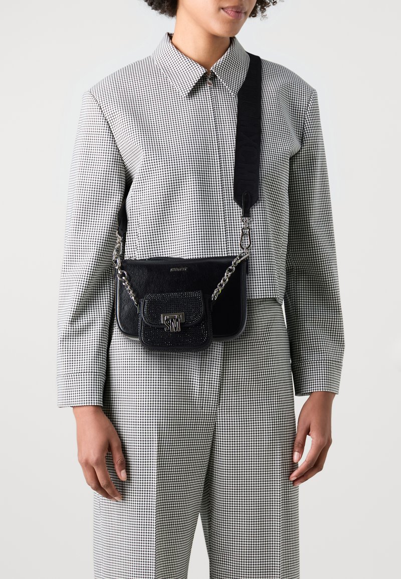 Borsa a tracolla nera in pelle con finitura testurizzata, dettagli in catena e una piccola tasca frontale. Indossata con una camicia a scacchi e pantaloni coordinati.