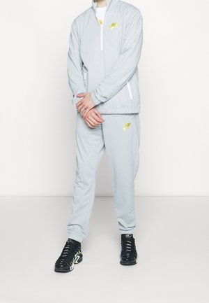 Osoba ubrana w jasnoszary dres Nike z żółtymi logo i czarne trampki stoi na białym tle.