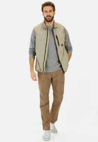 Beige ärmellose Weste mit grauem Pullover darunter, kombiniert mit hellbraunen Hosen und weißen Sneakers. Verfügt über eine Reißverschlusstasche an der Weste.