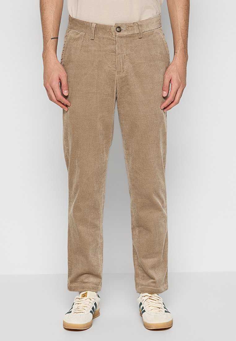 Beige Cordhosen mit geraden Beinen, die eine vertikale Rippstruktur, einen Knopfverschluss und Taschen aufweisen. Tragen mit weißen Turnschuhen.