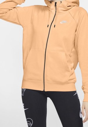 Personne portant un sweat à capuche zippé beige clair avec logo Nike et des leggings noirs ornés de plusieurs logos Nike blancs et de textes.