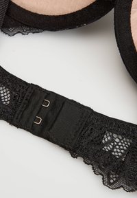 Čierny pásik na podprsenke z čipky s dvojitým zapínaním na háčiky a očká na svetlom povrchu, zobrazujúci detailný vzor čipky a prešitie.