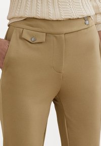 Béžové nohavice s prispôsobeným strihom a hladkou textúrou, s gombíkovým zapínaním na malej vrecku so záklopkou a bočnými vreckami, ktoré sú všité.