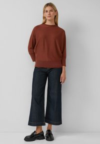Brązowy sweter z dzianiny z szerokim dekoltem, połączony z ciemnymi, szerokimi dżinsami i czarnymi lakierowanymi mokasynami, stojący na jasnym tle.