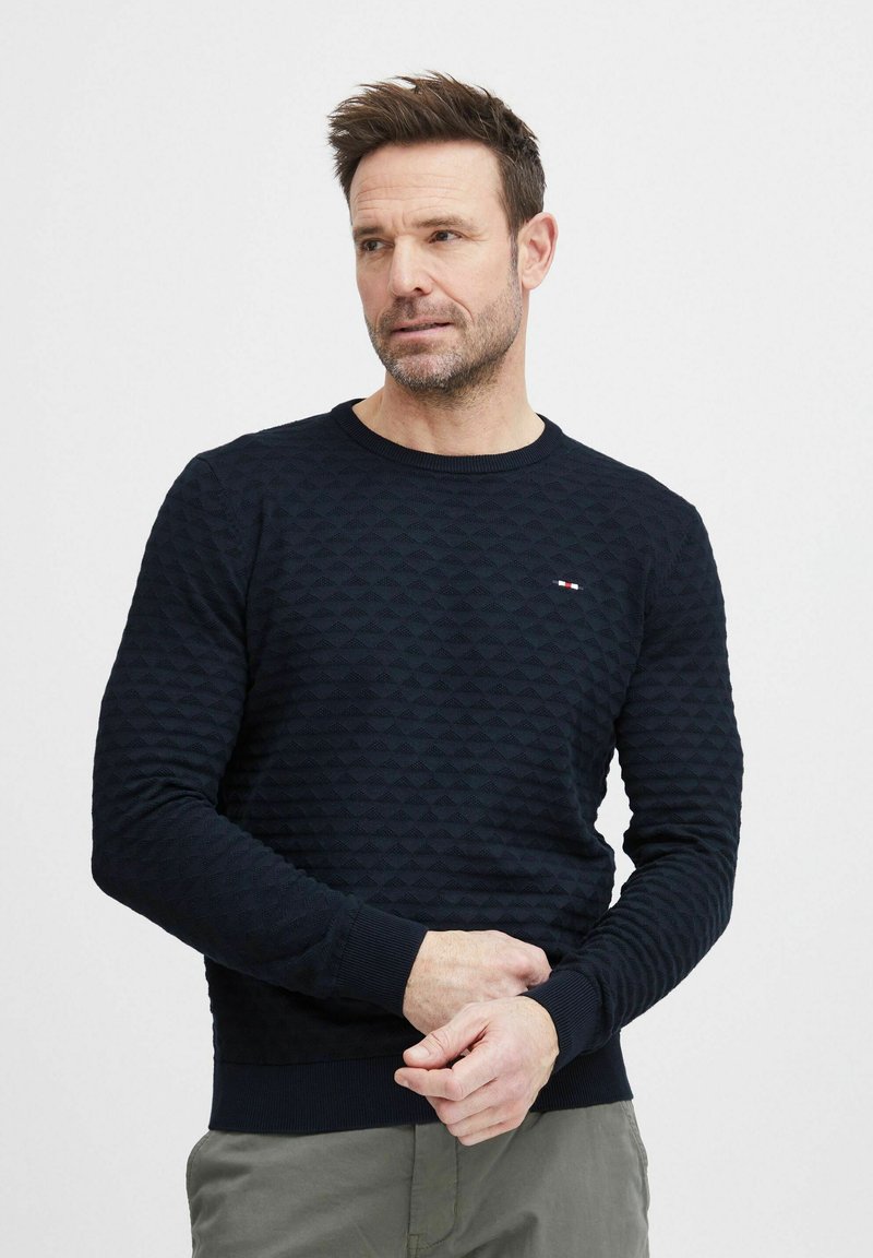 Granatowy sweter z teksturowanym wzorem diamentowym, okrągłym dekoltem i prążkowanymi mankietami oraz dołem. Dyskretna emblemat marki na piersi.