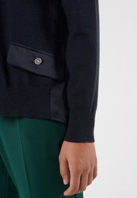 Pull en maille bleu marine avec poignets et poche côtelés, orné d'un détail de bouton argenté. Porté avec un pantalon vert émeraude, mettant en valeur le contraste des couleurs.