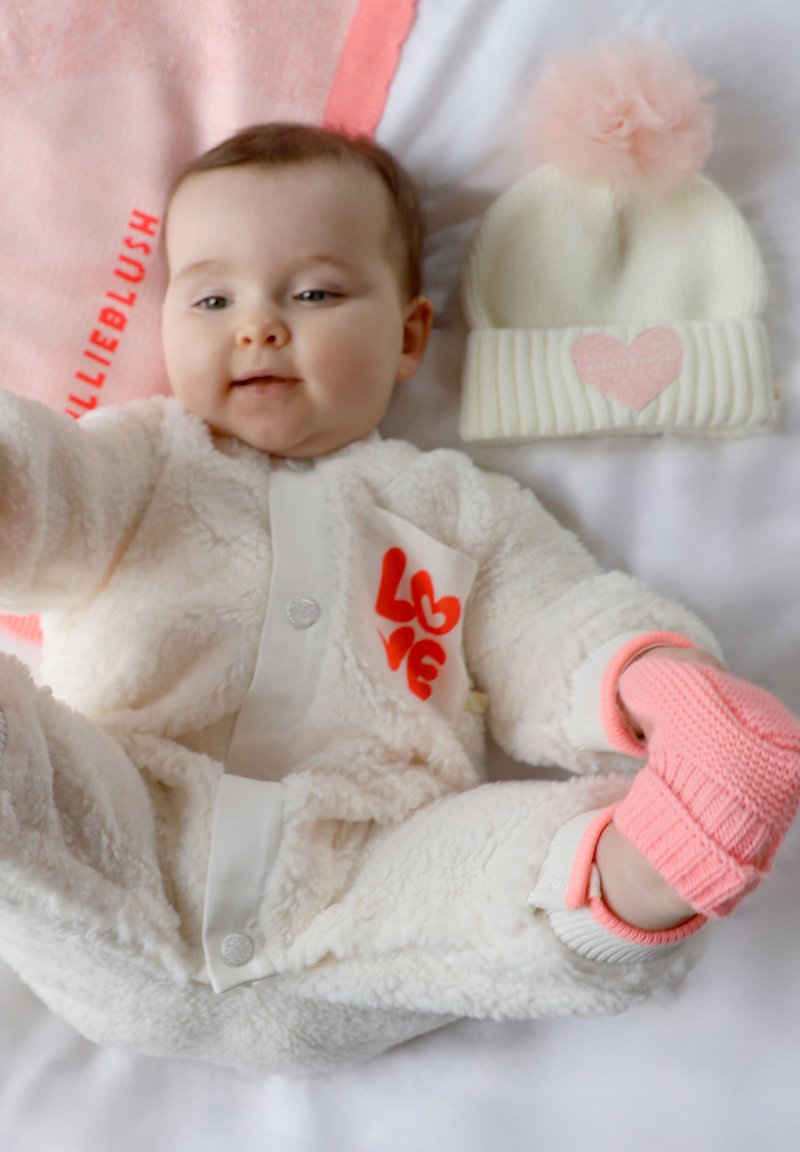 Romige fleece overall met rode "LOVE" detail, gecombineerd met een witte gebreide beanie met een roze pompon en hartaccent. Roze gebreide wanten.
