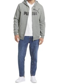 Felpa grigia con zip e logo Puma nero, indossata sopra una T-shirt bianca, abbinata a jeans blu e sneakers bianche.
