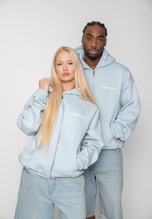 Zwei Modelle tragen hellblaue Kapuzenpullover mit dem Schriftzug "MULTIPLY" und stehen mit neutralen Gesichtsausdrücken nah beieinander vor einem einfarbigen Hintergrund.