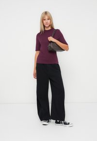 Femme portant un haut violet à manches courtes, un large pantalon noir et des baskets montantes noires, tenant un petit sac à main sombre sur son épaule.