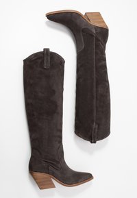 Botas de ante marrón oscuro hasta la rodilla con tacones de madera apilados, punteras puntiagudas y lengüetas de tirón en la parte superior sobre un fondo blanco.