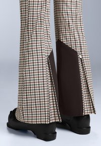 Nohavice s rozšírenými nohavicami v houndstooth vzore s hnedým panelom a zipsami na spodnej časti, skombinované s čiernou obuvou.