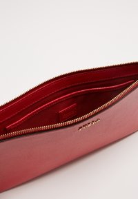 Clutch de cuero rojo con superficie texturizada, cremallera dorada y interior de satén. Incluye múltiples compartimentos para organización y un detalle con el logo.
