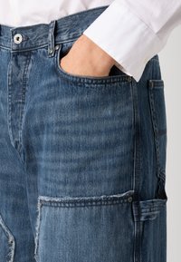Denim kavbojke srednje modre barve z šivano teksturo, opremljene s sprednjo žepno in srebrno gumbno podrobnostjo, z nagibi obrabljenosti.