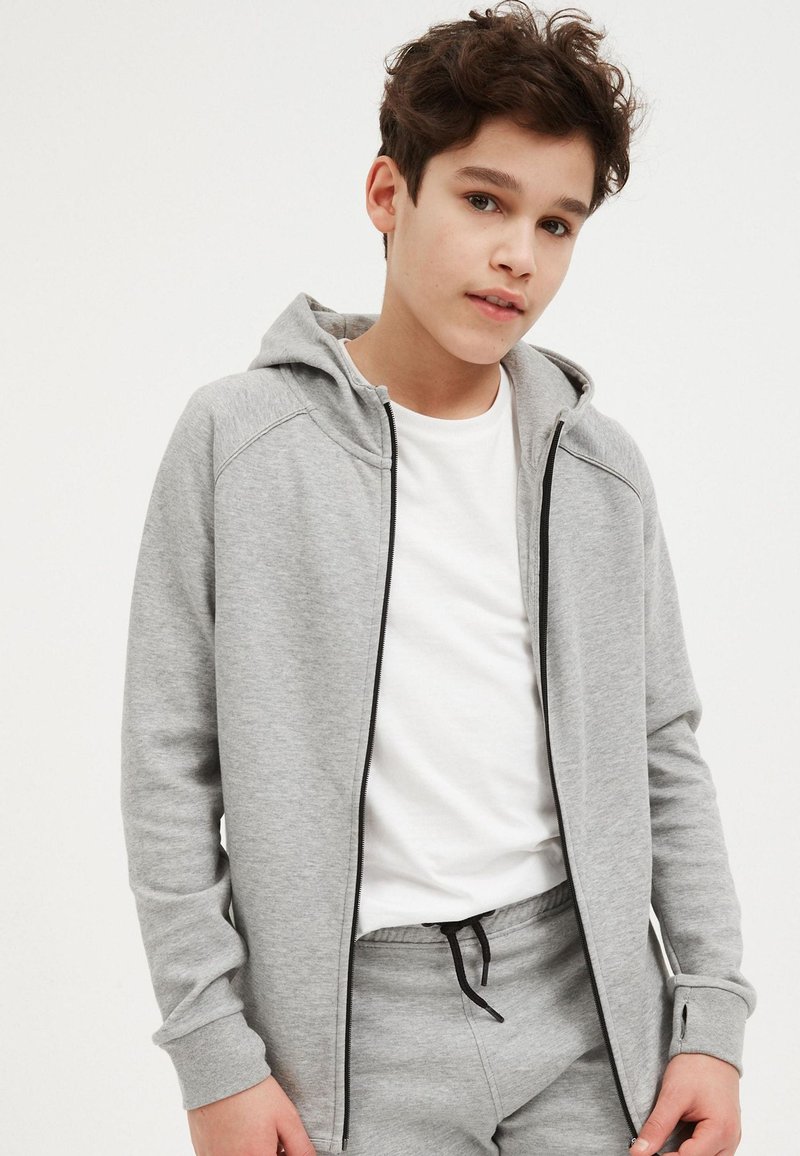 Adolescent portant un sweat à capuche gris zippé sur un T-shirt blanc et un pantalon de jogging gris assorti avec un cordon noir, debout devant un fond uni.