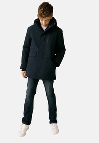 Marineblauer wasserdichter Parka mit Kapuze, ausgestattet mit Fronttaschen und einer Reißverschlusstasche auf der Brust, getragen über einem weißen Hemd, kombiniert mit dunklen Jeans und weißen Sneakers.