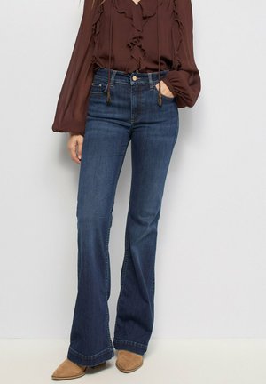 Mujer con vaqueros acampanados azul oscuro, blusa marrón con volantes y mangas largas, y zapatos de ante color canela con punta afilada, de pie contra un fondo claro.