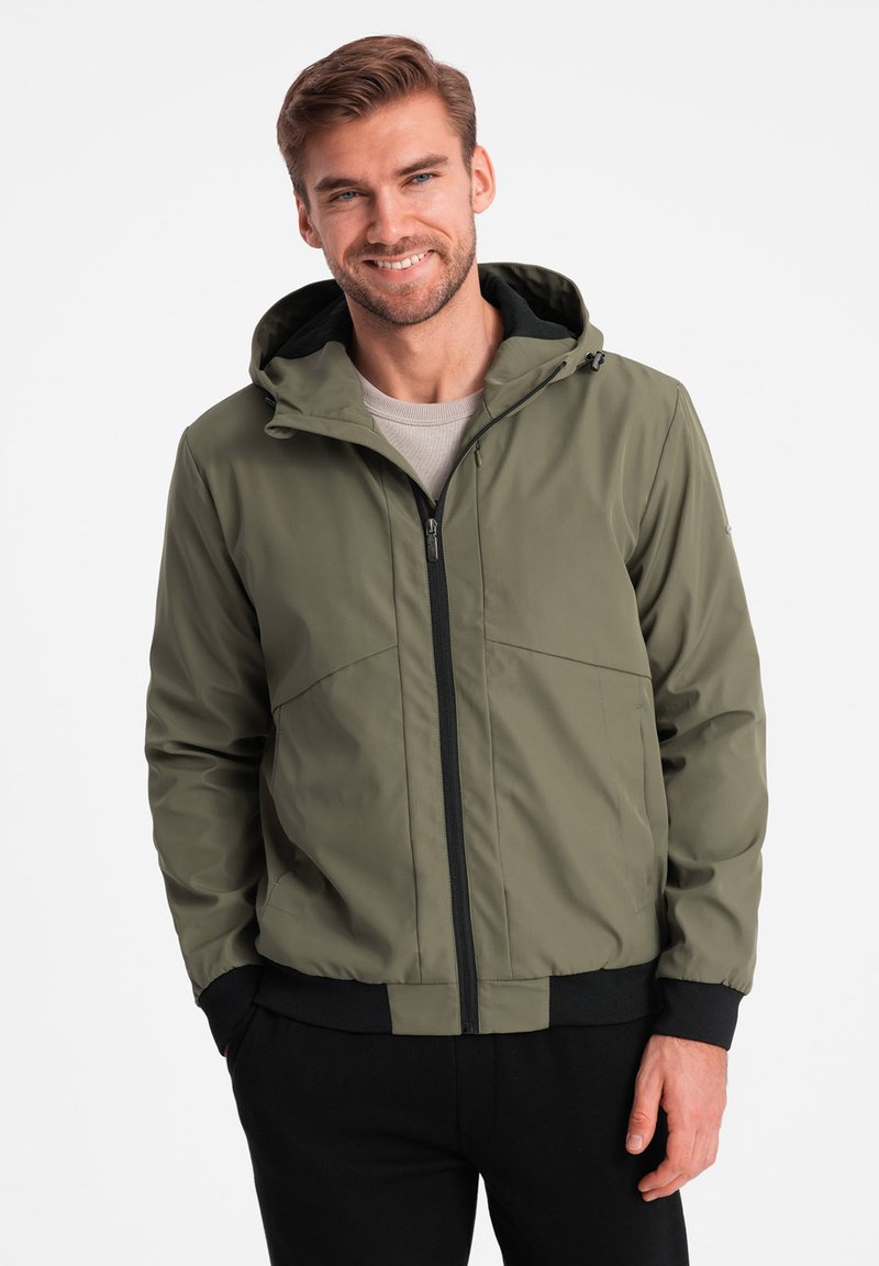 Giacca zip-up verde oliva con cappuccio, caratterizzata da una texture liscia, cerniera nera e polsini elastici. Indossata sopra una maglietta chiara.
