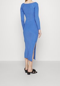 Långärmad, ribbad klänning i klarblått med sidslitsar. Har en bred rund halsringning och en figurnära silhuett, tillsammans med svarta klacksandaler.