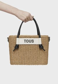 Torebka z plecionych słomek z czarnymi uchwytami z sztucznej skóry i białą taśmą z napisem "TOUS" w czarnym kolorze. Prostokątny kształt, chropowata powierzchnia.