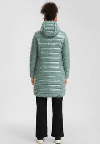 Abrigo largo acolchado en verde claro con acabado brillante, capucha y acolchado vertical. Lleva puesto sobre pantalones oscuros y zapatos informales.