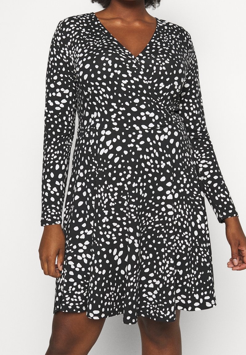 Czarna sukienka z długimi rękawami, z dekoltem w serek i wzorem białych nieregularnych plam. Materiał wydaje się gładki i lekko elastyczny.