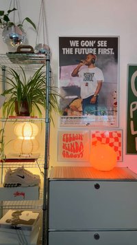 Metal shelving unit with white drawers, a wooden lamp, orange globe light, sneaker box, and posters with text. Green plant hanging above.