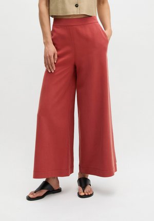 Person iført højtaljede, vidde rustfarvede bukser, beige cropped top og sorte åbne sandaler, står med hænderne i lommerne.