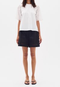 Bílý bavlněný tričko s krajkovými rukávy, doplněné černými šortkami s výřezovými akcenty na lemu. Model nosí černé sandály.
