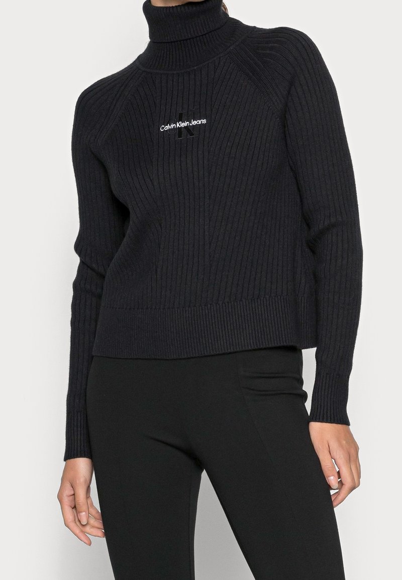 Pull en tricot côtelé bleu marine foncé avec col haut, arborant un logo sur la poitrine. Manches longues et coupe courte avec une finition texturée.