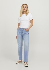 T-shirt bianca in cotone abbinata a jeans denim azzurri chiari, caratterizzati da una vestibilità comoda e un design a cinque tasche, completati da sneakers marroni.