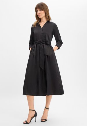 Frau trägt ein schwarzes Midi-Hemdkleid mit dreiviertel Ärmeln, einem gebundenen Taillengürtel und schwarzen High Heels mit Knöchelriemen vor weißem Hintergrund.