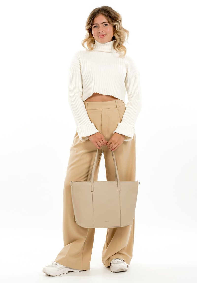 Young woman wearing a white cropped turtleneck sweater and beige wide-leg trousers, holding a large beige tote bag and white sneakers.