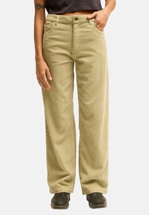 Personne portant un pantalon beige en velours côtelé à jambes larges, des bottes noires et un top noir court, debout avec la main dans la poche sur fond blanc.