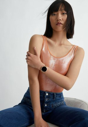 Roze gouden metallic bodysuit met dunne banden, gecombineerd met hoge blauwdenim jeans met knoopaccenten en een armbandhorloge met een zwarte wijzerplaat.